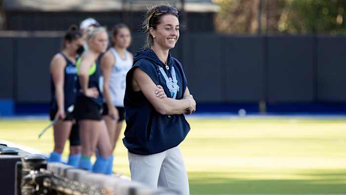 UNC field hockey coach Erin Matson smiles while watching practice with players standing behind her.